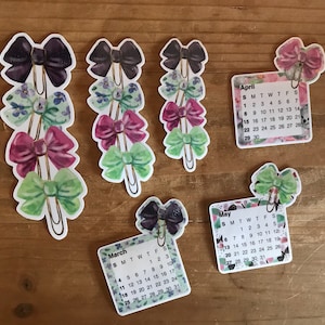 May include: Five colorful bow-shaped paper clips with floral designs. Each clip is attached to a calendar card with the months of March, April, and May. The calendar cards have a floral design and list the days of the week and dates.