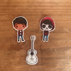 May include: Three stickers featuring characters and a guitar from the animated movie Coco. The characters are depicted in a cartoon style, with one wearing a red hoodie and skull face paint. The guitar is white with decorative details.