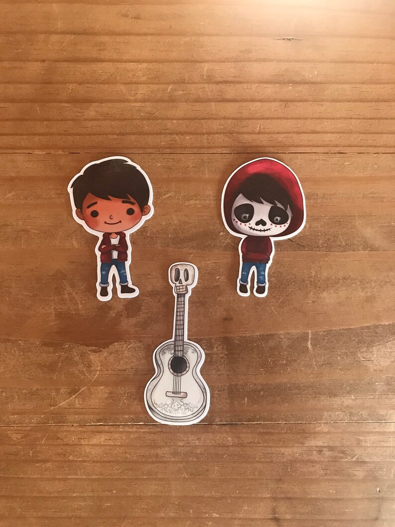May include: Three stickers featuring characters and a guitar from the animated movie Coco. The characters are depicted in a cartoon style, with one wearing a red hoodie and skull face paint. The guitar is white with decorative details.