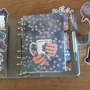 May include: A gray and pink planner with a galaxy design and gold rings. The planner has a sticker on the front that says "Magic Spells" and depicts a hand holding a white mug. The planner is surrounded by various stickers, including a crystal, a moon, an eye, and a purse.