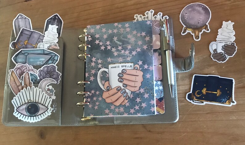 May include: A gray and pink planner with a galaxy design and gold rings. The planner has a sticker on the front that says "Magic Spells" and depicts a hand holding a white mug. The planner is surrounded by various stickers, including a crystal, a moon, an eye, and a purse.