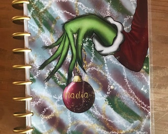 May include: A green Grinch hand holding a red Christmas ornament with the name "Nadia" written on it. The background is a blue and green swirl pattern with gold glitter.