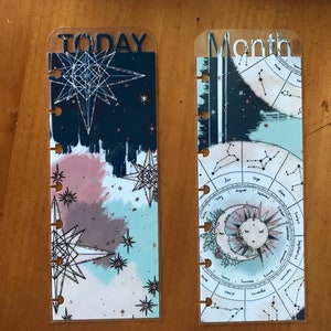 May include: Two clear plastic bookmarks with a celestial theme. The "TODAY" bookmark features a blue and pink watercolor background with gold glitter and a large geometric star. The "Month" bookmark features a circular zodiac chart with constellations and a crescent moon and sun.