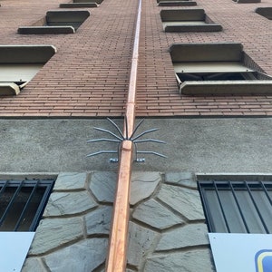 May include: A copper downspout with a metal bird deterrent attached to the bottom. The downspout is attached to a brick building with a stone facade.