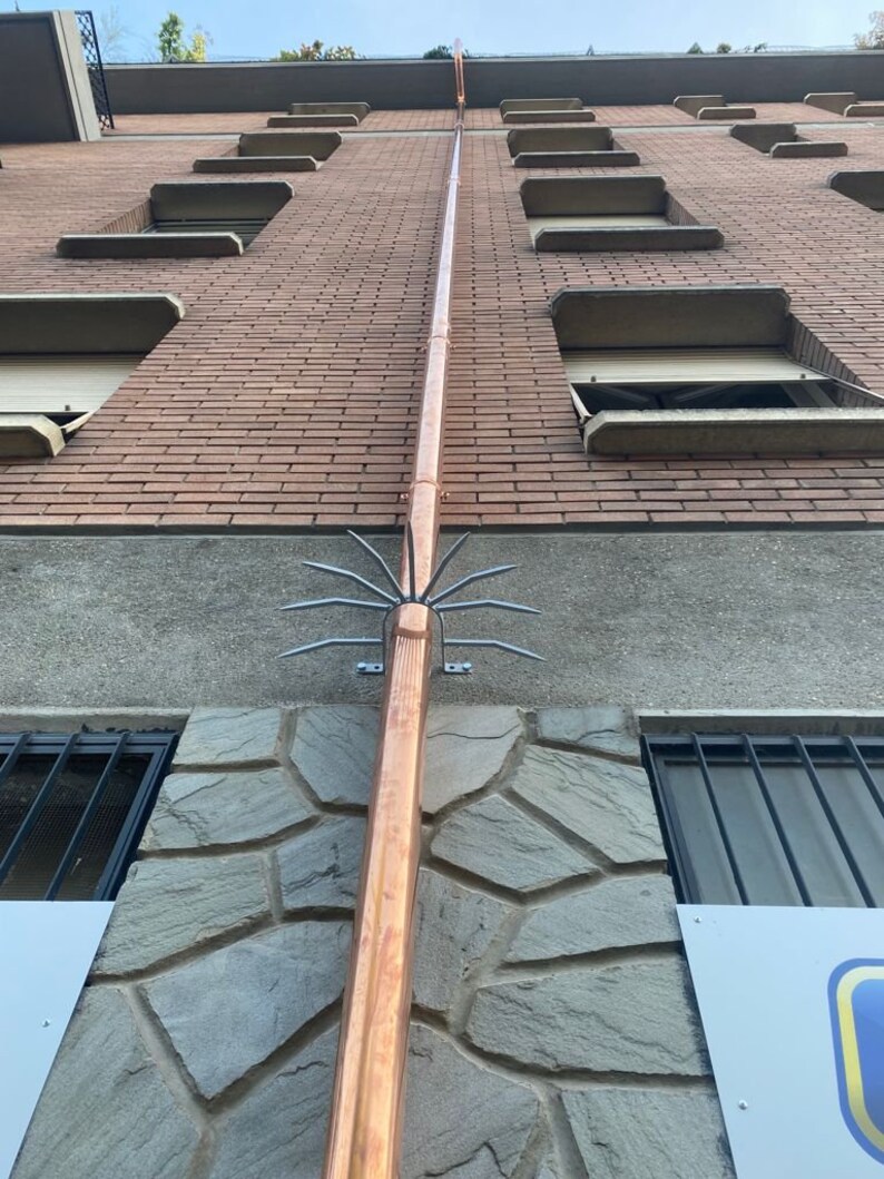 May include: A copper downspout with a metal bird deterrent attached to the bottom. The downspout is attached to a brick building with a stone facade.