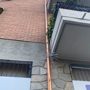 May include: A copper downspout with a metal bird deterrent attached to the side of a brick building. The downspout is attached to a stone wall with a white sign.