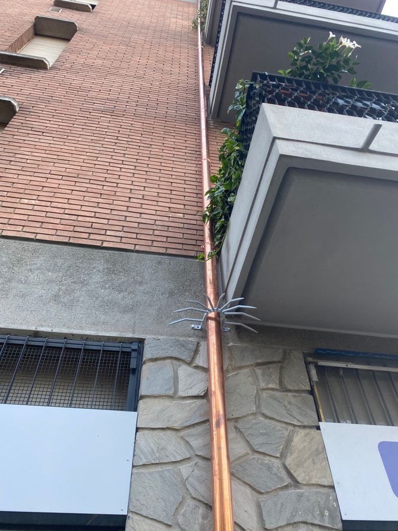 May include: A copper downspout with a metal bird deterrent attached to the side of a brick building. The downspout is attached to a stone wall with a white sign.