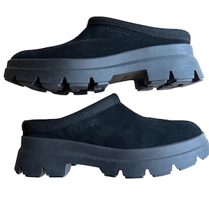 May include: A pair of black slip-on clogs featuring a soft suede upper and a thick, chunky rubber lug sole. The shoes have a rounded toe design and a debossed logo on the side of the upper, providing a solid black aesthetic with a heavy tread.