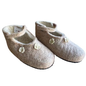 May include: A pair of beige felt slippers with a Mary Jane strap and small flower accents. The slippers have a soft, textured appearance and a light-colored interior. The soles are dark, and the overall design is simple and cozy.