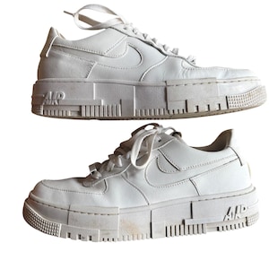 May include: Two white leather sneakers with thick, pixelated soles. The Nike swoosh is visible on the sides. The word "AIR" is printed on the sole near the heel. The shoes have white laces.