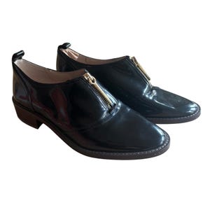May include: Pair of black patent leather shoes with a gold zipper detail. The shoes have a low heel and a rounded toe. The zipper runs down the center of the shoe. The shoes are on a white background.
