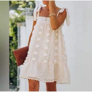 May include: Off-white, knee-length dress with a square neckline and tie straps. The dress features a textured, dotted pattern and a ruffled hem. A brown clutch is held in the arm.