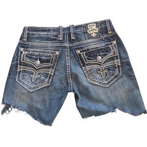 May include: Blue denim shorts with distressed edges. The shorts feature decorative stitching, embellished back pockets with a fleur-de-lis design, and a leather patch with the text "Rock Revival".