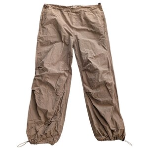 May include: Light brown trousers featuring a relaxed fit with a gathered elastic waistband and adjustable drawstring hems at the ankles. The pants are constructed from a lightweight, slightly wrinkled fabric and include side pockets for utility.