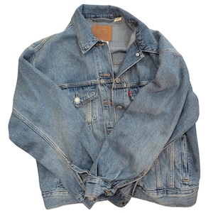 May include: A light blue denim jacket with a classic design. The jacket features a button-up front, two chest pockets, and two side pockets. A brown leather patch is visible on the back of the collar. The jacket has a red tag on the left chest pocket.