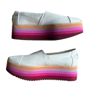 May include: Pair of white canvas platform slip-on shoes with a multi-colored striped sole. The sole features layers of orange, pink, and red hues. The shoes have a rounded toe and a simple, casual design.