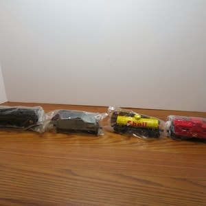 May include: A set of four model train cars individually wrapped in clear plastic. The collection includes a black locomotive, a silver Shell tanker, a yellow Shell tank car with red lettering, and a red caboose, all arranged in a horizontal row.