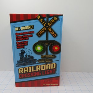 May include: A box for a railroad crossing light replica. The box is blue with red and white stripes. The front of the box features a picture of a train and the words "Railroad Crossing Light". The box also includes the words "All Aboard!" and "Red and Green Lights".