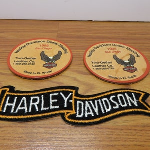 May include: Two brown leather Harley-Davidson Dealer Meeting coasters with an eagle and Harley-Davidson logo. The coasters are made by Two-Gether Leather Co. and say "Made in Ft. Worth". A black and orange embroidered Harley-Davidson banner patch.