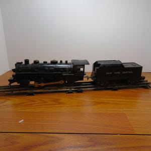 May include: A black toy train set with a locomotive and a separate car on a track. The locomotive has a detailed design with various components. The car is labeled "NEW YORK CENTRAL". The set is placed on a wooden surface.