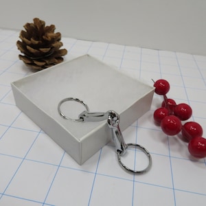 May include: A silver-toned key chain with two rings and a decorative horseshoe-shaped charm. The key chain is displayed on a white surface with a pine cone and red berries in the background.