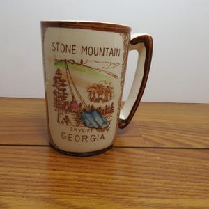 May include: Cream-colored ceramic mug with a brown handle and rim. The mug features a scenic illustration of Stone Mountain, Georgia, with the text "STONE MOUNTAIN" and "GEORGIA". The illustration includes a skylift and trees.