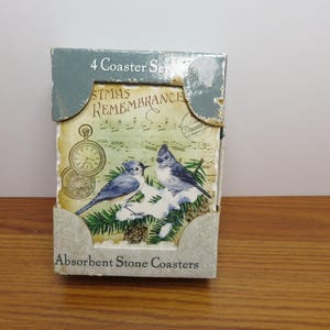 May include: A set of four absorbent stone coasters in a cardboard display box. The box features the text 4 Coaster Set, Christmas Remembrance, and Absorbent Stone Coasters. The coasters depict two blue birds on pine branches with a pocket watch.