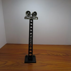 May include: A black metal model of a tall light fixture with two circular lights at the top. The fixture has a series of oval cutouts along its length and sits on a square base. The lights are silver-colored.