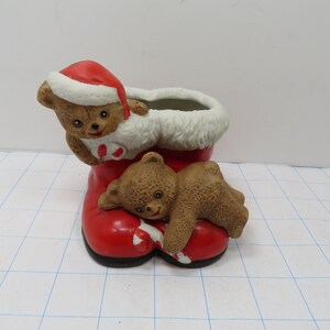 May include: A ceramic Christmas boot planter with two brown teddy bears. The boot is red with white fur trim and a red Santa hat. The bears are holding candy canes.