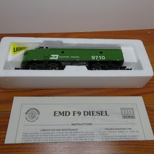 May include: A green and gray model train locomotive, marked with the number 9710, sits in a white foam container. The model is labeled "Burlington Northern." The packaging includes a paper with the text "EMD F9 DIESEL" and "HO SCALE."