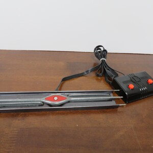 May include: A vintage model train track section with a red and silver decorative center. A black control box with two red buttons is connected to the track via a black wire. The track is on a brown surface.