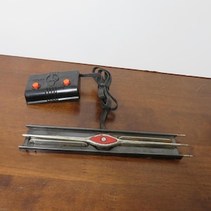 May include: A vintage model train track section with a black control box featuring two orange buttons. The track has a red and silver center detail. A black cord connects the track and control box.