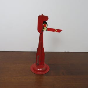 May include: A vintage red metal toy train signal with a round base and a tall, vertical body. The signal arm extends horizontally, featuring red and yellow accents. The toy is designed to stand upright.