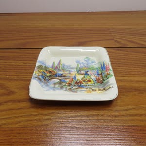 May include: A small, square, off-white dish with a painted landscape scene. The scene depicts a garden with a bridge, trees, and a figure in a dress. The dish has a gold trim and the words "Old World Garden".