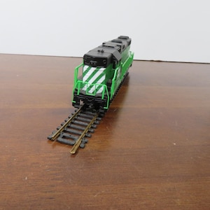 May include: A green and black model train locomotive with white diagonal stripes on the front. The model train is on a section of black and gold-colored train tracks. The train is on a brown surface.