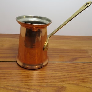 May include: A copper coffee pot with a wide, flared rim and a long, angled brass handle. The interior of the pot is lined with a silver-toned metal. The exterior surface is smooth and polished, reflecting light across its cylindrical body.