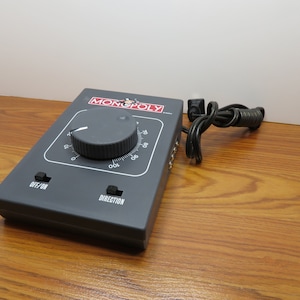May include: A gray Monopoly electric train controller with a black knob, an on/off switch, and a direction switch. The controller has a black power cord.
