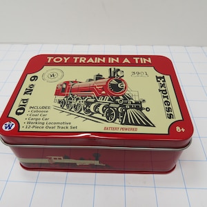 May include: A red rectangular tin toy train set box with the words "TOY TRAIN IN A TIN" and "Express" printed on it. The box features a red and black illustration of a steam train and includes a caboose, coal car, cargo car, working locomotive, and a 12-piece oval track set.
