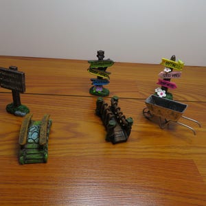 May include: A collection of miniature garden decorations, including a signpost, a bridge, a wheelbarrow, and directional signs. The items are made of resin and painted in various colors, with a weathered appearance. The signposts have text.
