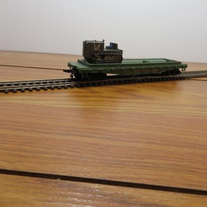 May include: A model train flatcar in olive green carrying a small, dark brown tracked bulldozer. The flatcar is positioned on a section of model train track with black ties and silver rails, featuring a detailed coupling mechanism at the end.
