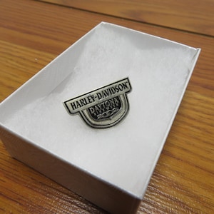 May include: A Harley-Davidson Daytona 200 pin in a white gift box. The pin is silver and black, with the Harley-Davidson logo and "Daytona 200" text. The box is lined with white fabric and sits on a wooden surface.