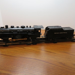 May include: Black toy train set with a locomotive and a boxcar. The locomotive has a gold-colored trim and the boxcar has the words "Penn Central" printed on the side.
