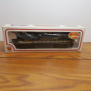 May include: A black and silver HO scale model train locomotive, labeled "SOUTHERN" and "7076", is displayed in a white box with a red border. The box also features "8 Wheel Drive" and "Bachmann" branding. The train is visible through a clear plastic window.