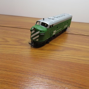 May include: A green and silver model train engine with a streamlined design. The front features diagonal white stripes. The train is on a wooden surface.