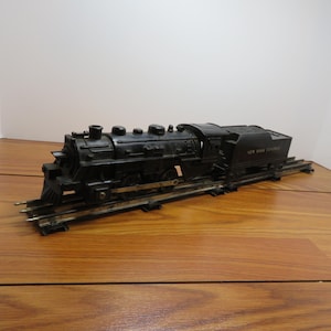 May include: A black model train on tracks, featuring a locomotive and a coal car. The train is set on a wooden surface. The coal car has the text "NEW YORK CENTRAL" printed on it.