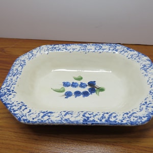 May include: A white ceramic serving dish with a blue and white speckled glaze. The dish has a rectangular shape with rounded corners and features a painted design of blue blueberries with green leaves.