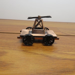 May include: A miniature copper-colored toy cart with four black wheels. The cart has a detailed frame and a propeller-like structure on top. The toy is set against a wooden surface.