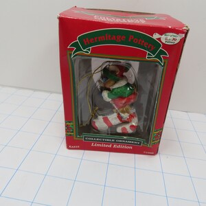 May include: A red box with a white and green Christmas ornament inside. The ornament features a bear wearing a green hat and red and white striped stocking. The box reads "Hermitage Pottery" and "Collectible Ornament Limited Edition".