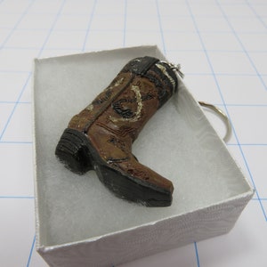 May include: A brown cowboy boot keychain with a silver horseshoe design and white stitching. The keychain is in a white box with a white background.