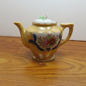 May include: A small, antique teapot with a yellow glaze and floral design. The teapot features a curved spout, a handle, and a lid with a small floral accent. The front of the teapot has a rectangular panel with a floral pattern in pink, red, and green.
